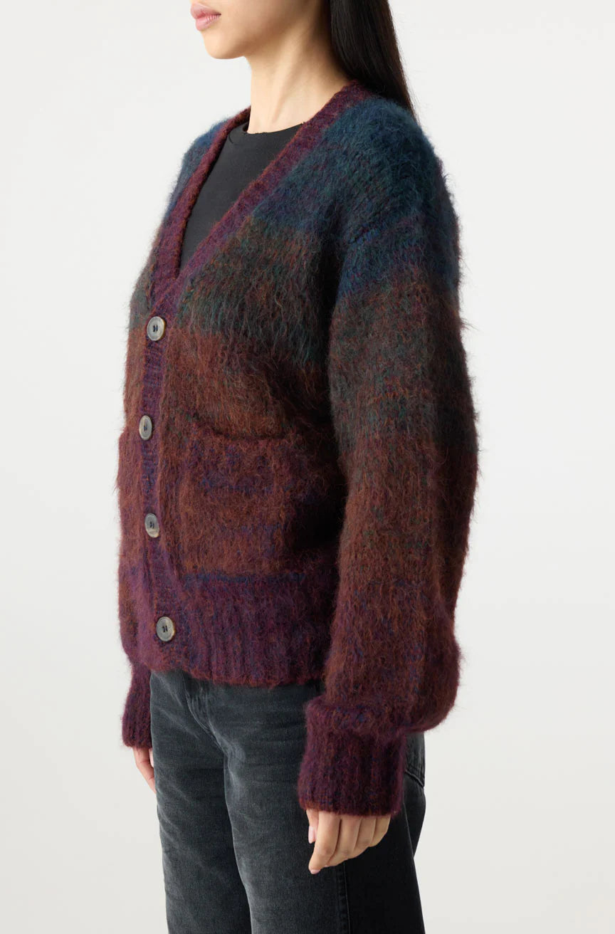 A person wears the Nancy Sweater by AMO Denim—a fuzzy, multicolored Suri Alpaca cardigan in purple, blue, and brown with large buttons and front pockets—over a black top and dark jeans, facing slightly left.
