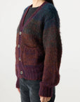 A person wears the Nancy Sweater by AMO Denim—a fuzzy, multicolored Suri Alpaca cardigan in purple, blue, and brown with large buttons and front pockets—over a black top and dark jeans, facing slightly left.