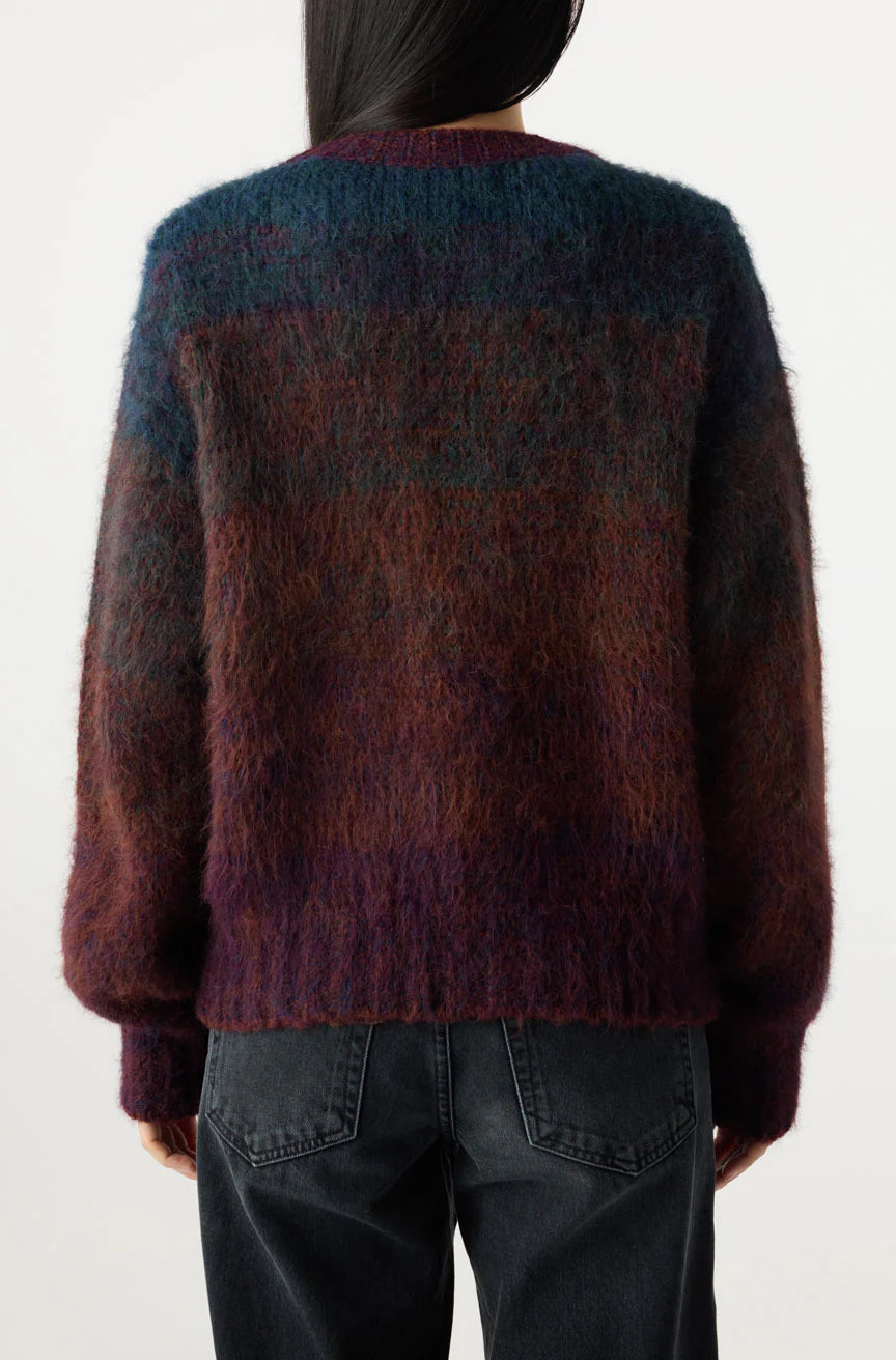 A person is seen from the back wearing the fuzzy, multicolored Nancy Sweater by AMO Denim in blue, purple, and brown, paired with dark gray jeans against a plain white background.