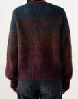 A person is seen from the back wearing the fuzzy, multicolored Nancy Sweater by AMO Denim in blue, purple, and brown, paired with dark gray jeans against a plain white background.
