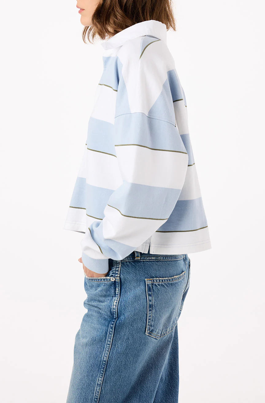 A person with shoulder-length hair wears the Crop Annie Rugby by AMO Denim in light blue and white stripes, paired with blue jeans. They stand sideways against a plain white background with one hand in their pocket.