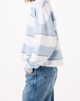 A person with shoulder-length hair wears the Crop Annie Rugby by AMO Denim in light blue and white stripes, paired with blue jeans. They stand sideways against a plain white background with one hand in their pocket.