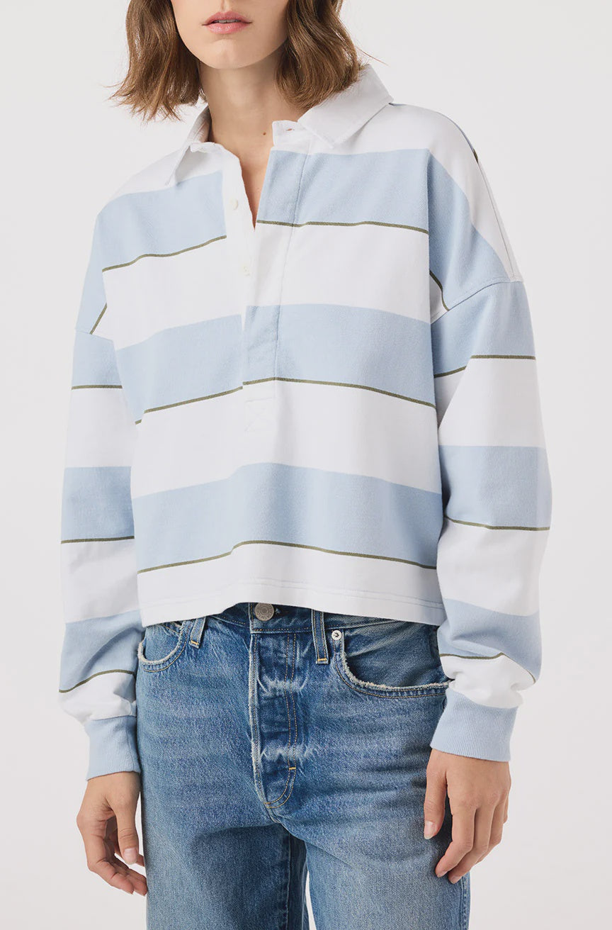 Someone is dressed in a Crop Annie Rugby by AMO Denim, featuring light blue and white stripes. This long-sleeved shirt pairs with blue jeans for a casual and relaxed look against a plain white background, with their face not visible.