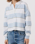 Someone is dressed in a Crop Annie Rugby by AMO Denim, featuring light blue and white stripes. This long-sleeved shirt pairs with blue jeans for a casual and relaxed look against a plain white background, with their face not visible.