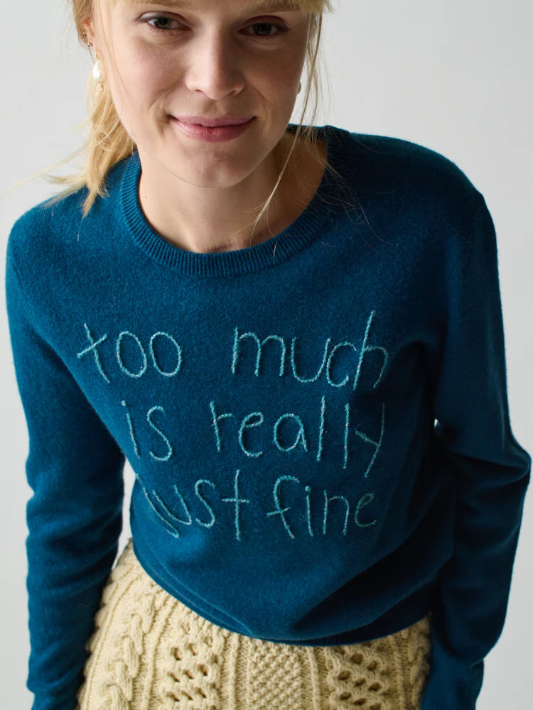 A blonde woman smiles softly in a Lingua Franca Cashmere Crew featuring handwritten-style "too much is really just fine" text and a beige knit skirt, set against a light, relaxed Montauk-inspired background.
