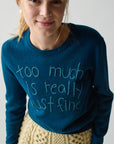 A blonde woman smiles softly in a Lingua Franca Cashmere Crew featuring handwritten-style "too much is really just fine" text and a beige knit skirt, set against a light, relaxed Montauk-inspired background.