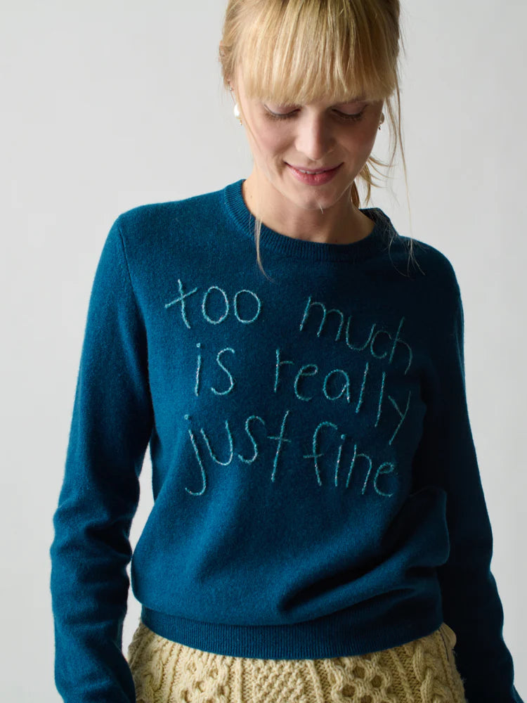 A woman with blonde hair and bangs smiles while wearing Lingua Franca’s Cashmere Crew in Montauk blue, embroidered with "too much is really just fine," paired with a cream textured skirt.