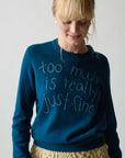 A woman with blonde hair and bangs smiles while wearing Lingua Franca’s Cashmere Crew in Montauk blue, embroidered with "too much is really just fine," paired with a cream textured skirt.