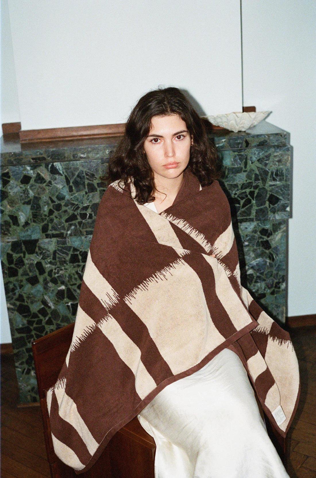 A woman with wavy dark hair sits on a chair, wrapped in the Autumn Sonata Karin Bath Sheet. She wears a white skirt and looks at the camera, with a dark green stone fireplace and white bowl visible in the background.