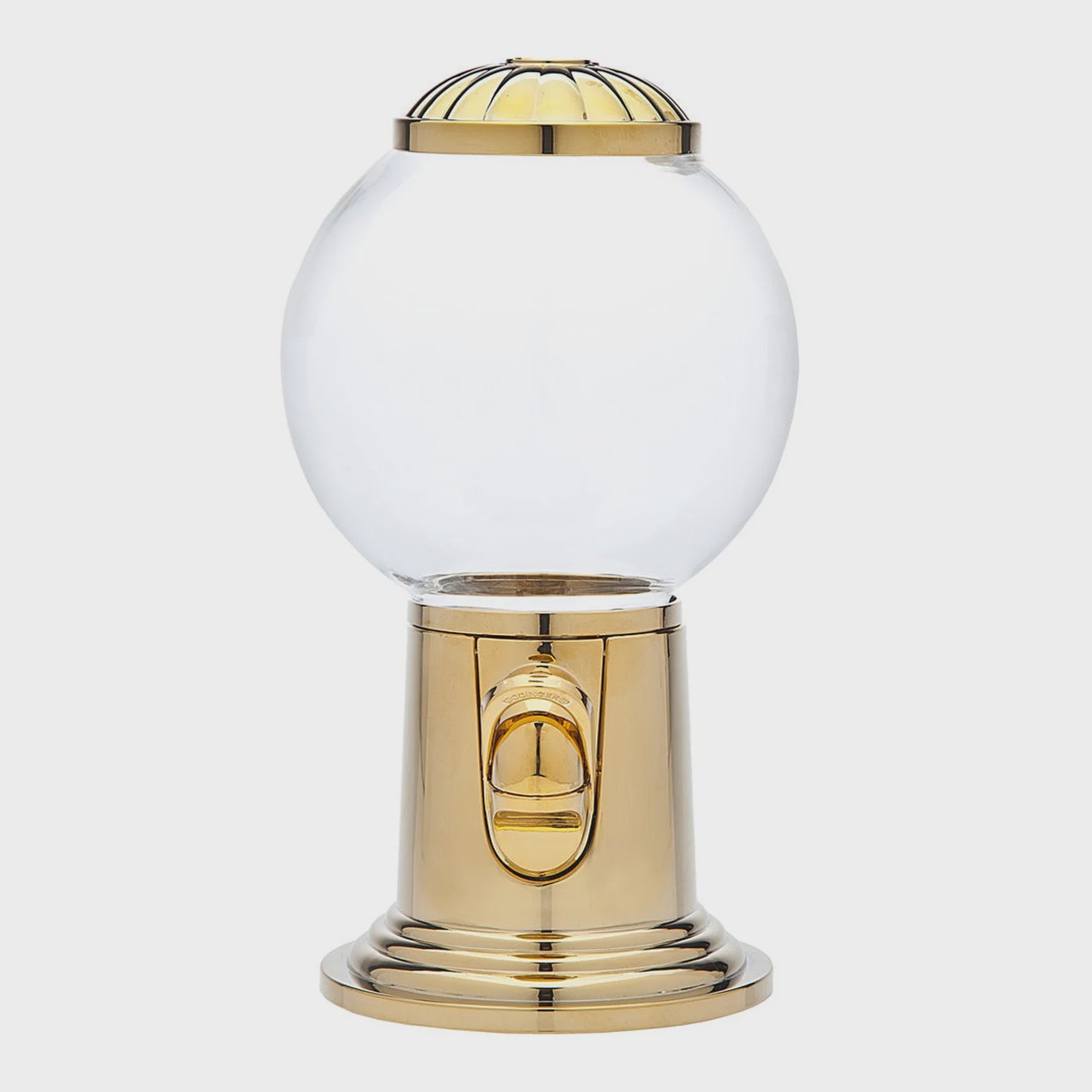 The Godinger (Faire) Candy Machine is a gold-toned candy dispenser that boasts a clear, round glass globe at the top and a gold lever for dispensing at the front. Its base features durable construction and complements the gold accents on the globe&#39;s top. This nostalgic piece is showcased against a plain white background.