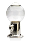 The Godinger (Faire) Candy Machine features a silver metallic base and lid, coupled with a transparent glass globe. The front boasts a coin slot and turn knob. This elegant candy dispenser, showcasing a timeless design, is displayed on a reflective white surface with no contents inside.