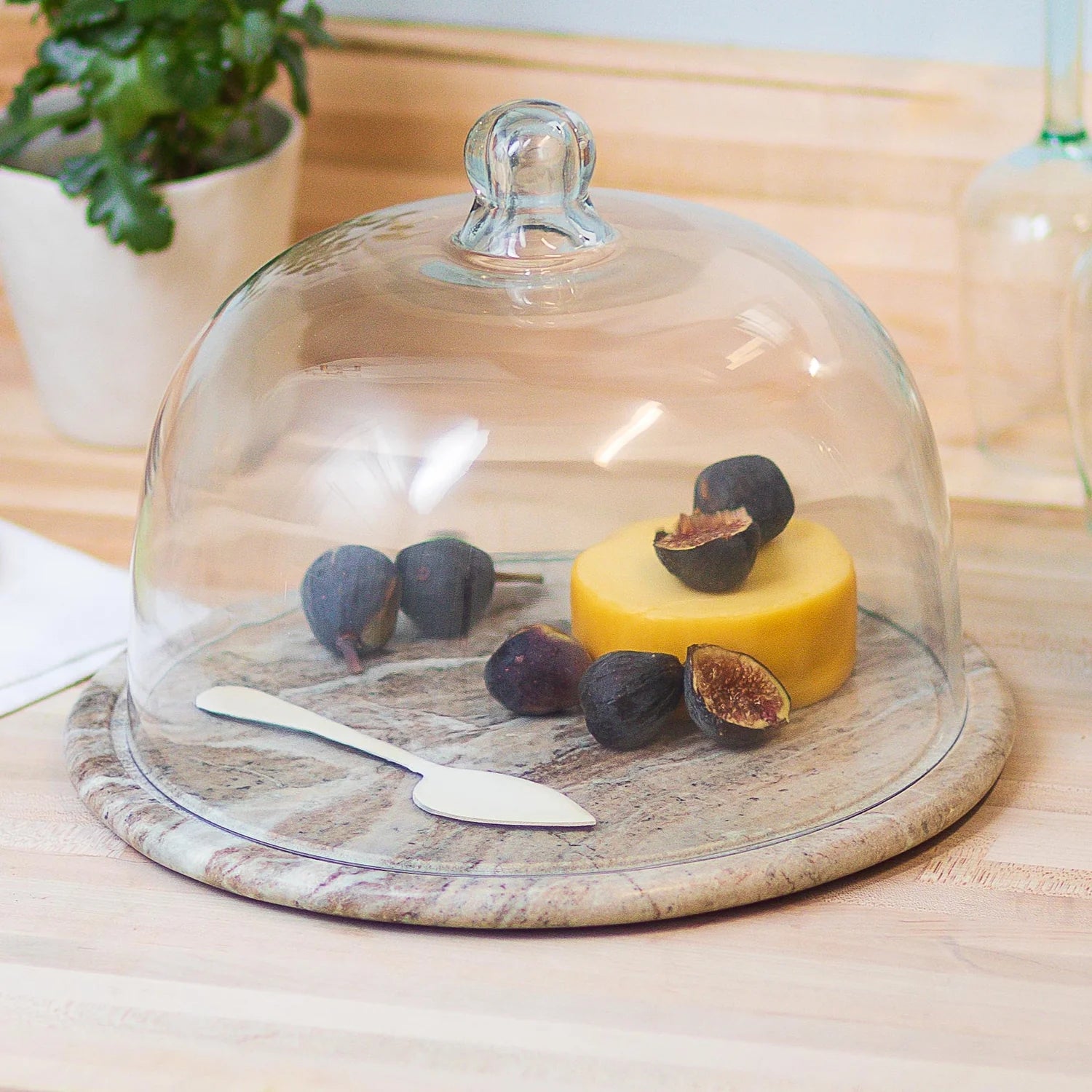 The Be Home Waterfall Marble Cloche features a round pink and teal marble cheese board with a glass dome, showcasing yellow cheese and figs with a spreader for stunning artisanal decor on your wooden countertop.