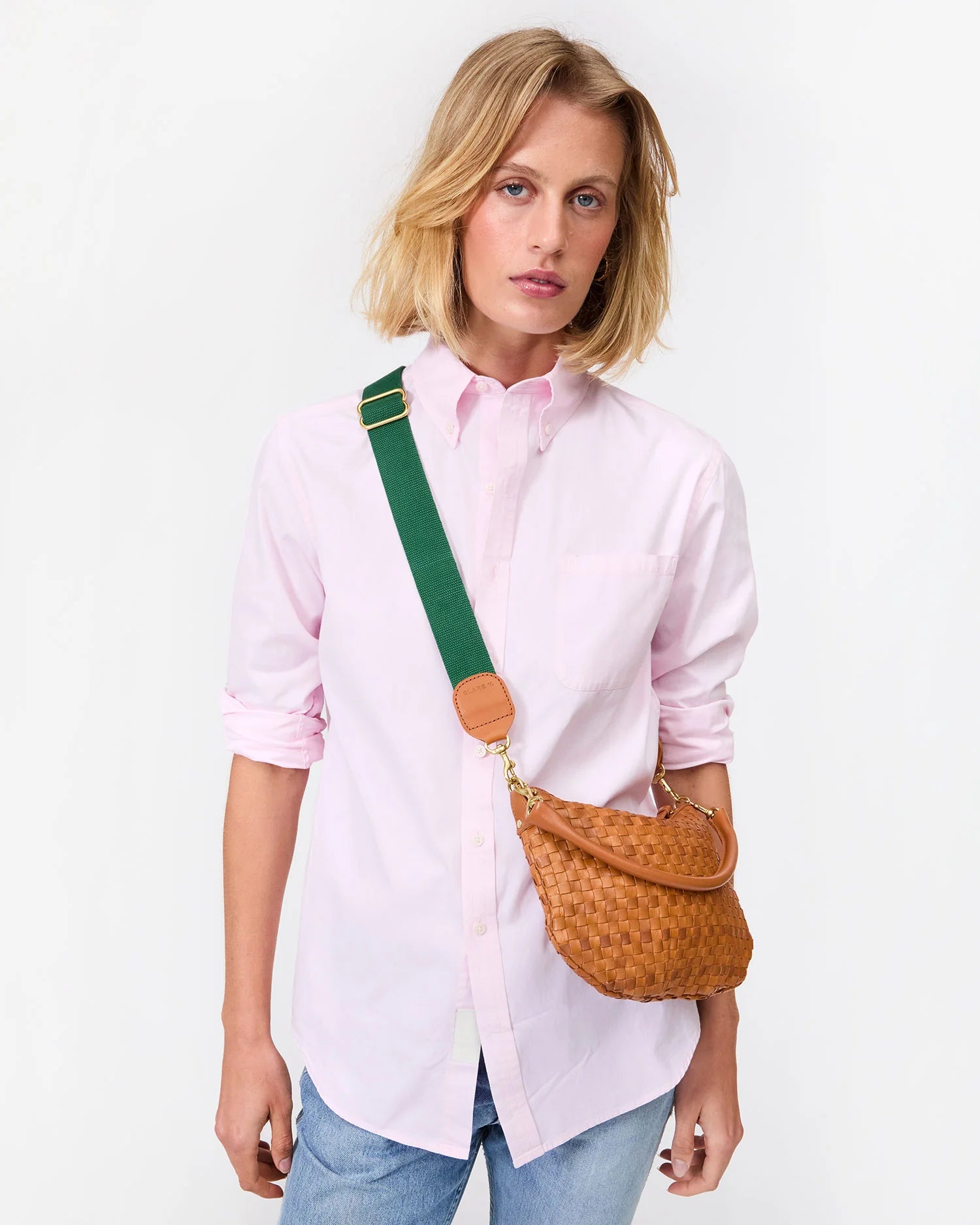 A person wearing a light pink button-up and blue jeans poses with the Clare Vivier Adjustable Crossbody Strap, a woven brown bag with a thick green cotton webbing strap, against a plain white background.