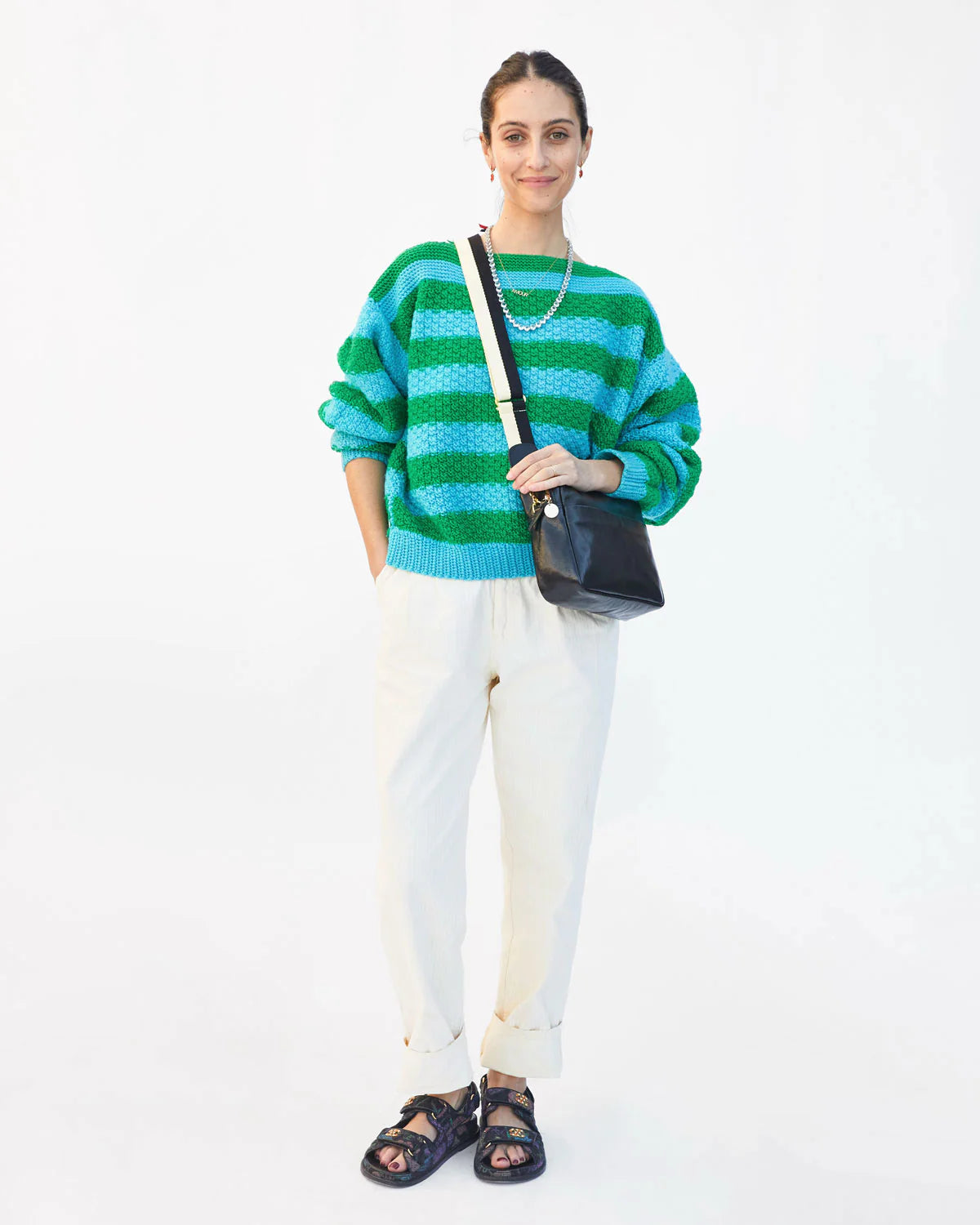A woman stands against a white background with her hand in her pocket. She is wearing a green and blue striped sweater, white pants, black sandals, and a black crossbody bag with a Clare Vivier Crossbody Strap. Her hair is tied back, and she is smiling.