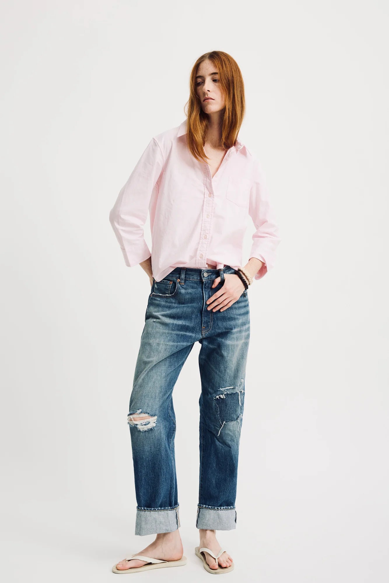 A person with long red hair is wearing a light pink button-up shirt and rolled-up Lucy Boyfriend Jean by Denimist. Barefoot, they exude a relaxed fit against the plain white background.