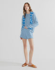 A woman poses against a white background wearing Kule’s The Agnes blue and white mini stripe long-sleeve shirt with matching cotton knit shorts, an elastic waistband, a blue sweater over her shoulders, and tan sandals.