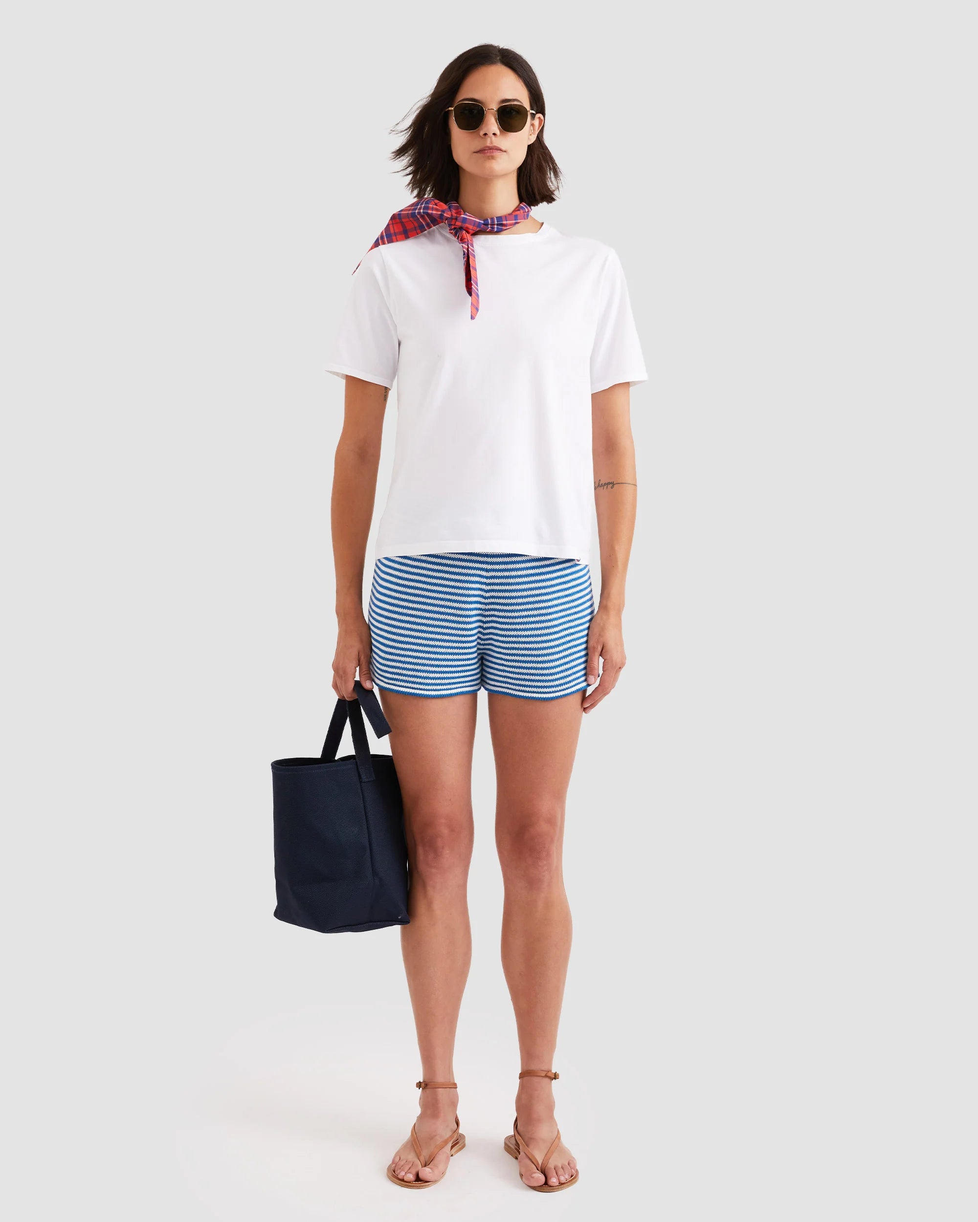 A woman wearing Kule’s The Agnes cotton knit mini-stripe shorts, a white tee, sunglasses, sandals, a red neck scarf, and holding a navy tote stands against a light background.