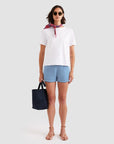 A woman wearing Kule’s The Agnes cotton knit mini-stripe shorts, a white tee, sunglasses, sandals, a red neck scarf, and holding a navy tote stands against a light background.