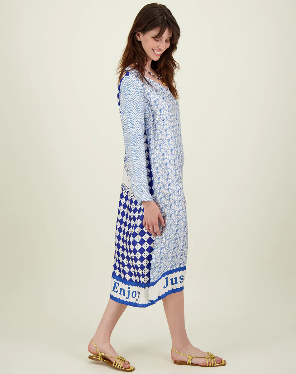 A woman with long brown hair, wearing the Me369 Violet V-Neck Tee Dress in blue and white with a geometric print and gold sandals, stands sideways and smiles. The dress hem features the words "Enjoy" and "Just.