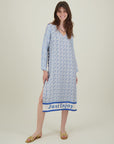 A woman smiles brightly in the Me369 Violet V-Neck Tee Dress, a loose long-sleeve T-dress with a blue and white geometric print and "Just Enjoy" at the hem, paired with yellow sandals and a necklace against a plain background.