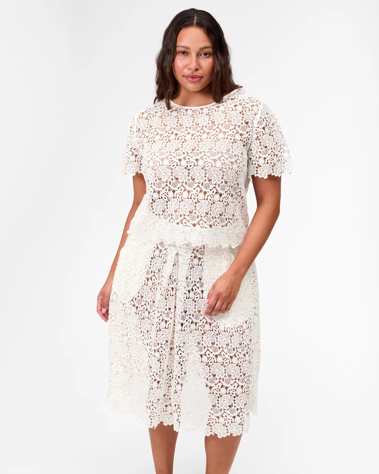 A woman stands against a plain white background wearing the Clare Vivier Lou Lou Top, a short-sleeved, white cotton lace top with floral pattern and see-thru design.