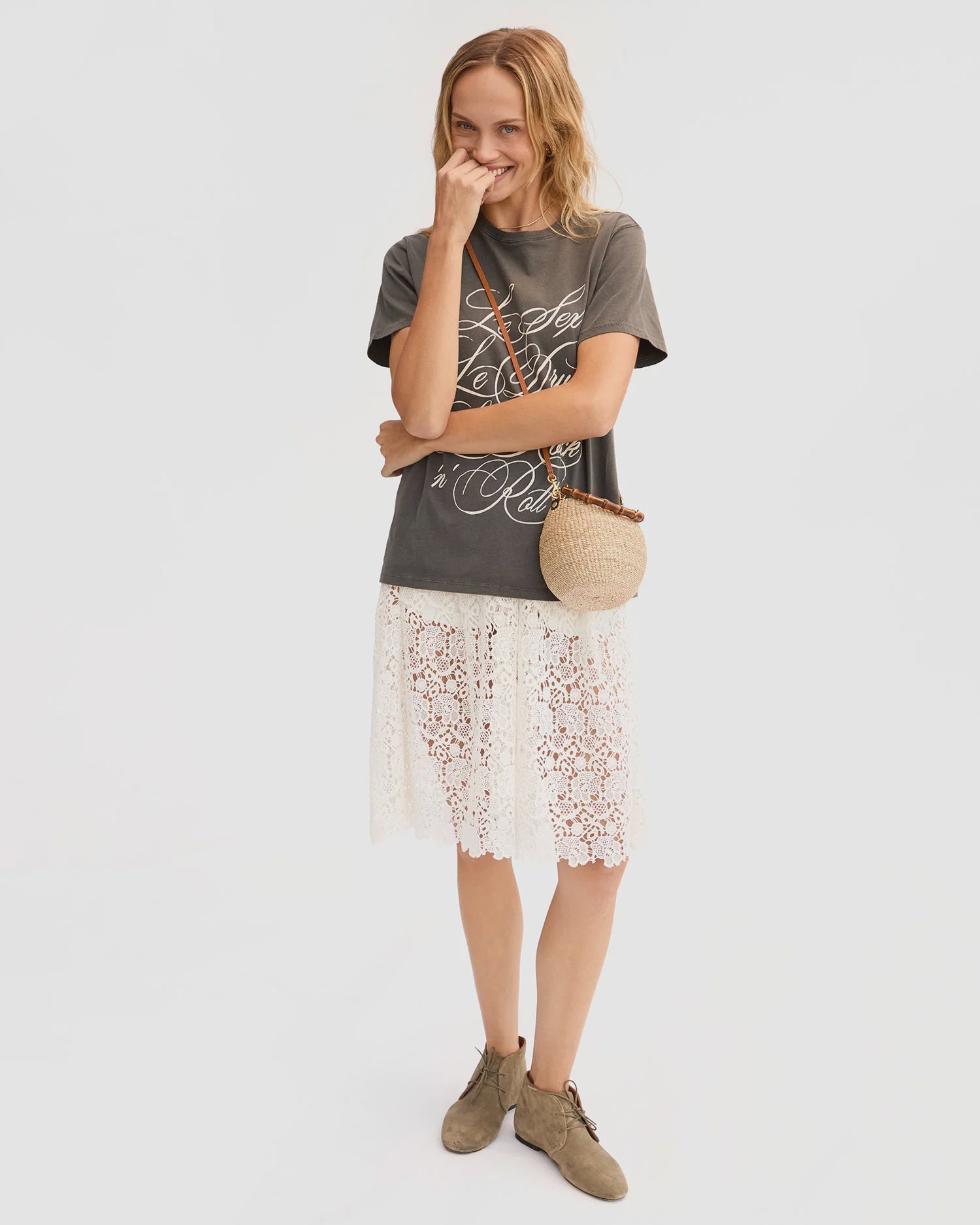A smiling woman stands against a white background wearing the Clare Vivier Anais Court Skirt, a gray graphic t-shirt, tan ankle boots, and a round woven crossbody bag, touching her face with one hand.