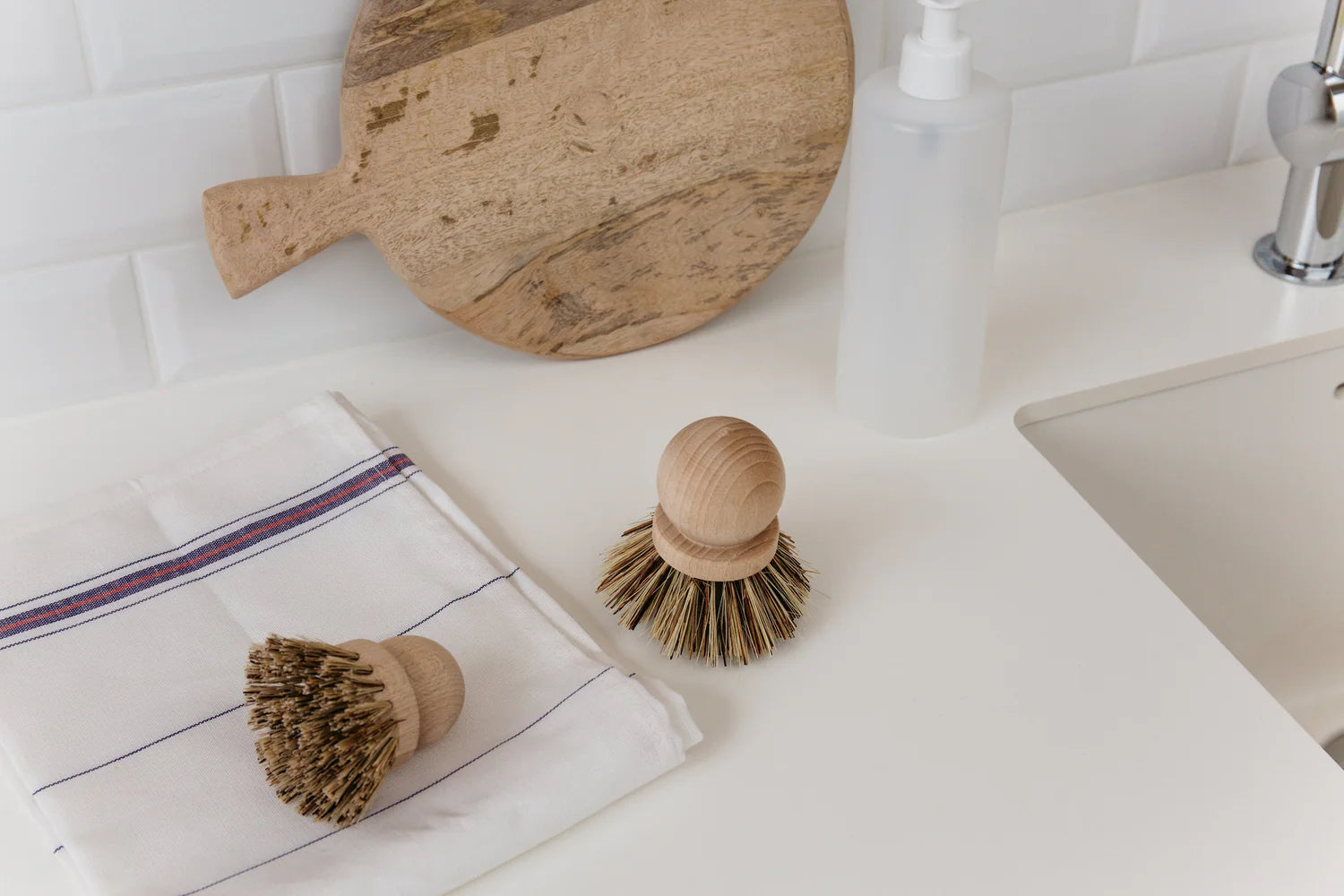 Two Be Home Saucepan Brushes with vegetable fiber bristles rest on a white countertop beside a folded towel, round cutting board, and soap dispenser near the kitchen sink.