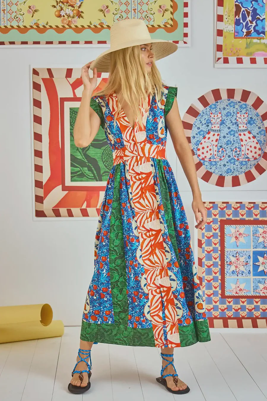 The woman wears a Hunter Bell NYC April Dress from the spring collection, showcasing green, blue, and Orange Grove mixed prints. She pairs it with a wide-brimmed hat and strappy sandals amidst brightly patterned framed artworks on the walls.