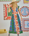 The woman wears a Hunter Bell NYC April Dress from the spring collection, showcasing green, blue, and Orange Grove mixed prints. She pairs it with a wide-brimmed hat and strappy sandals amidst brightly patterned framed artworks on the walls.