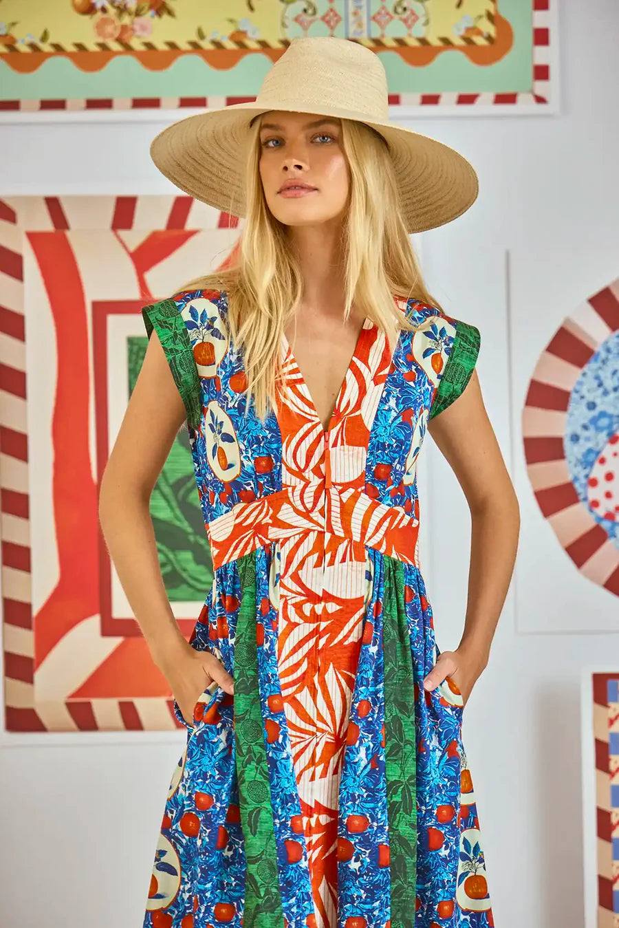 A woman with long blonde hair wears Hunter Bell NYC's April Dress, a V-neckline design with an Orange Grove mixed print in blue, red, orange, and green. She poses with hands in pockets against a colorful, patterned wall backdrop.