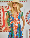 A woman with long blonde hair wears Hunter Bell NYC's April Dress, a V-neckline design with an Orange Grove mixed print in blue, red, orange, and green. She poses with hands in pockets against a colorful, patterned wall backdrop.