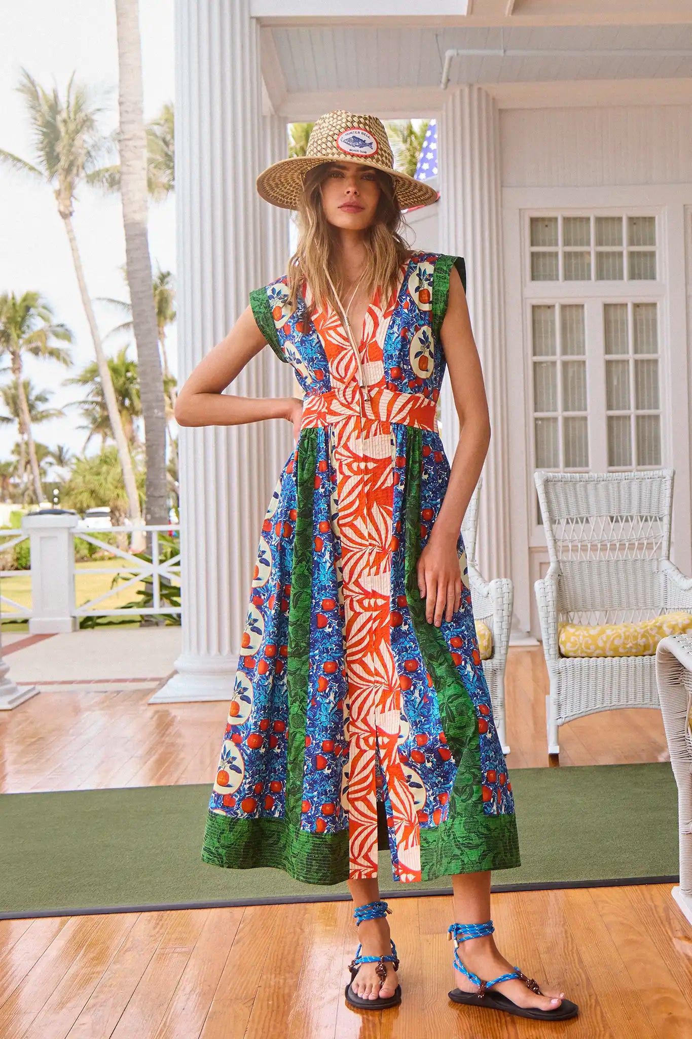 A woman in a Hunter Bell NYC April Dress featuring an Orange Grove mixed print and a straw hat stands on a porch, barefoot with blue sandals nearby. Wicker furniture and palm trees surround her, enhancing the tropical vibe.