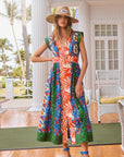 A woman in a Hunter Bell NYC April Dress featuring an Orange Grove mixed print and a straw hat stands on a porch, barefoot with blue sandals nearby. Wicker furniture and palm trees surround her, enhancing the tropical vibe.