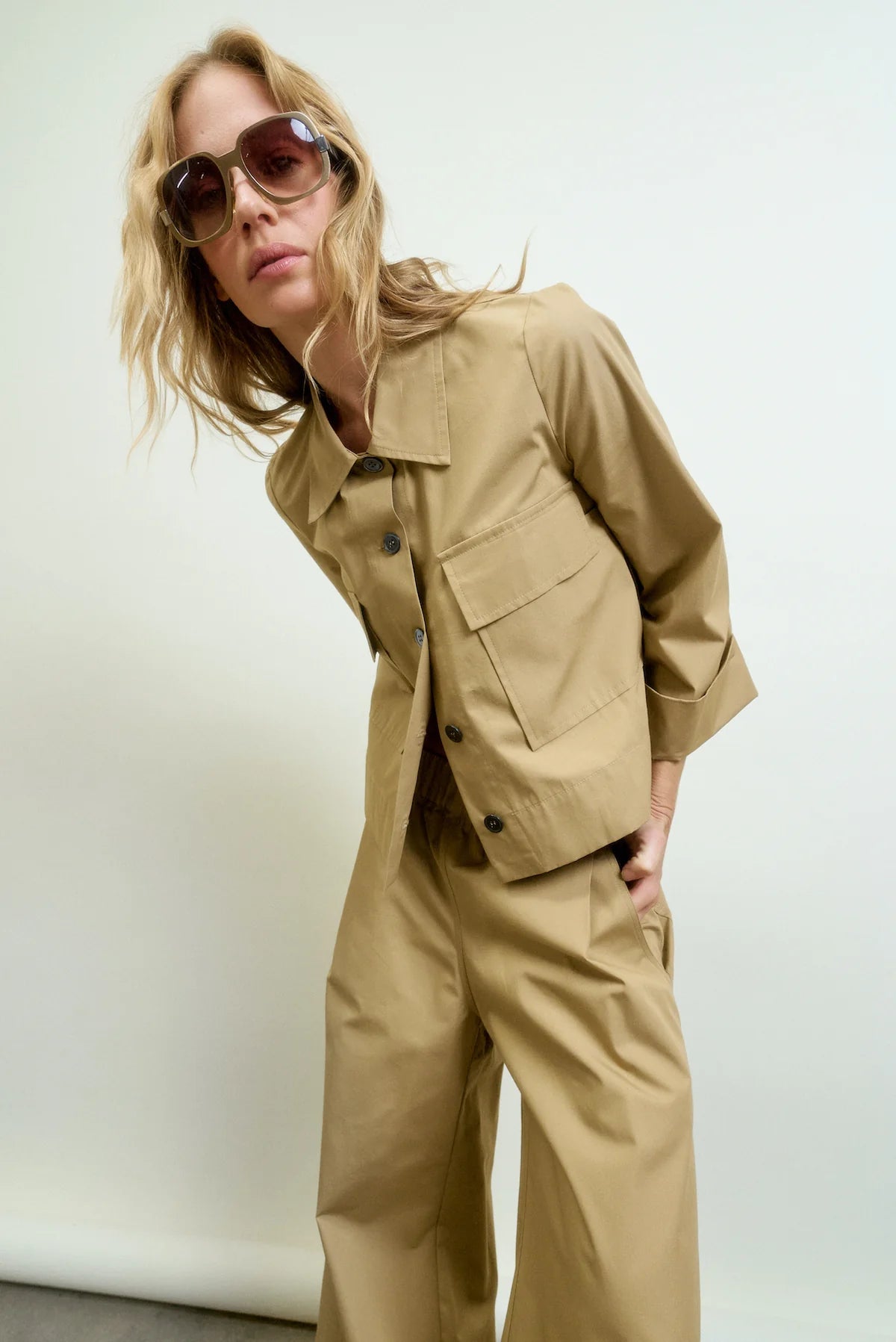 A person with wavy blonde hair wears oversized sunglasses and the Aquarius Cocktail Yes Sir Cropped Jacket with matching wide-leg pants, posing against a plain light background and leaning forward for a relaxed look.