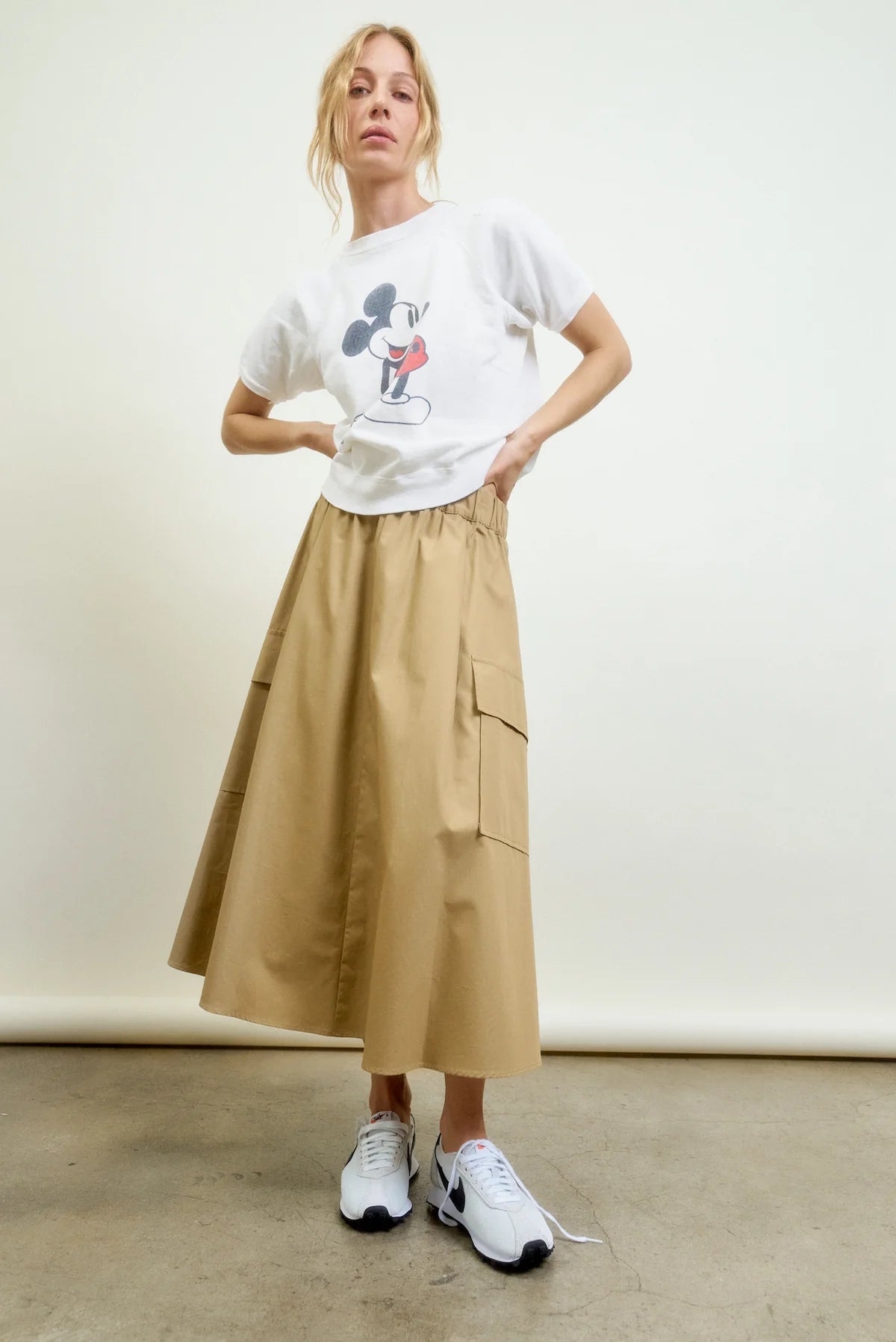 A woman stands against a plain background wearing a white Mickey Mouse t-shirt, white sneakers, and the Aquarius Cocktail Le Cargo Skirt—a tan Japanese cotton poplin skirt with oversized pockets. She poses relaxedly with one hand on her hip.