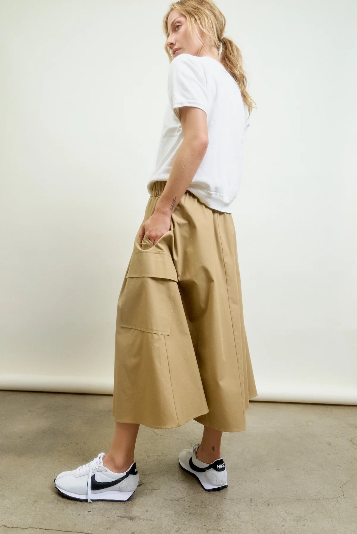 Someone stands sideways on a concrete floor against a plain light background, wearing a white t-shirt, white Nike sneakers, and the Le Cargo Skirt by Aquarius Cocktail—tan with oversized pockets in Japanese cotton poplin.