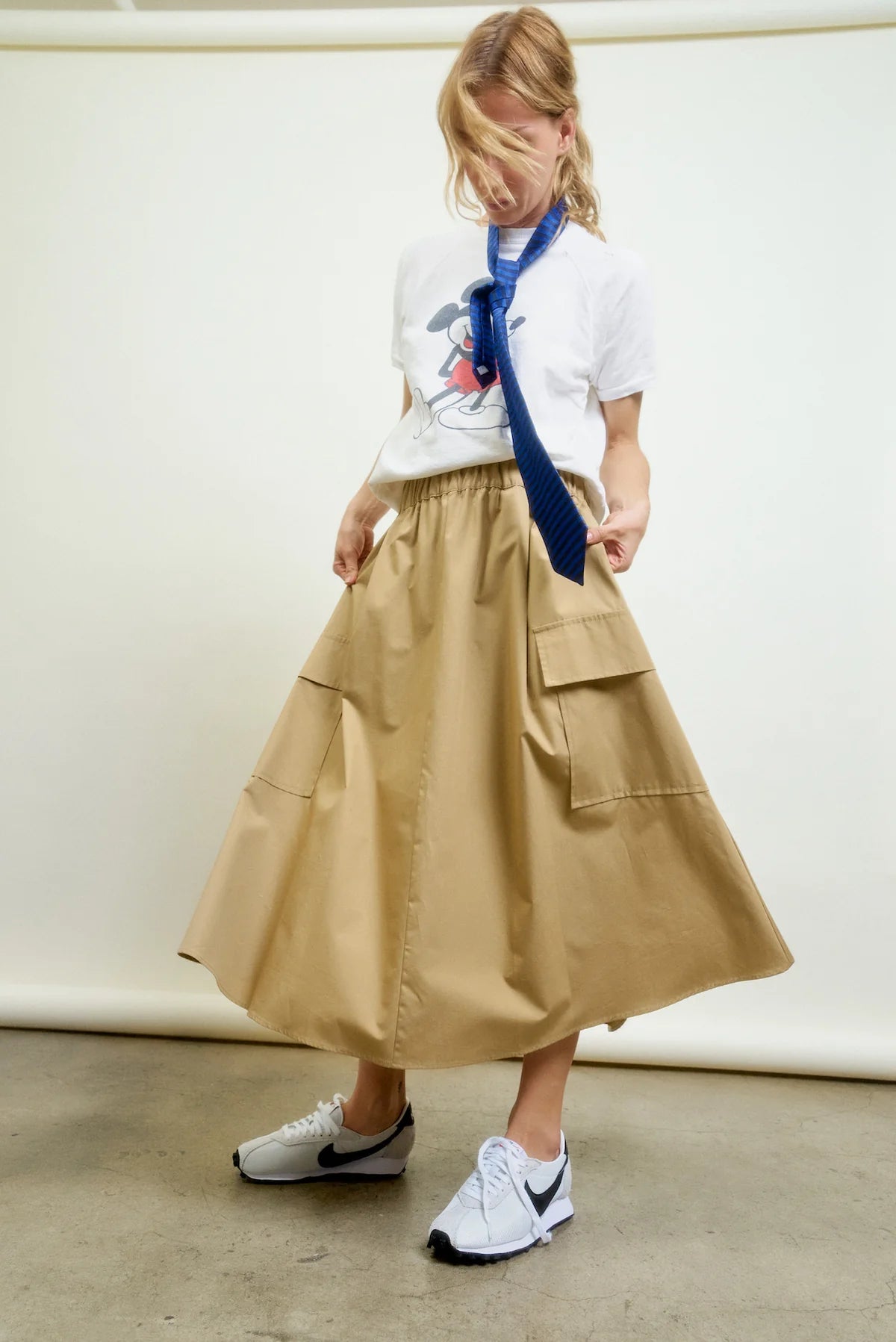 A person in a white Mickey Mouse t-shirt, blue tie, Le Cargo Skirt by Aquarius Cocktail with oversized pockets, and white Nike sneakers stands against a plain backdrop, looking down with hair partially covering their face.