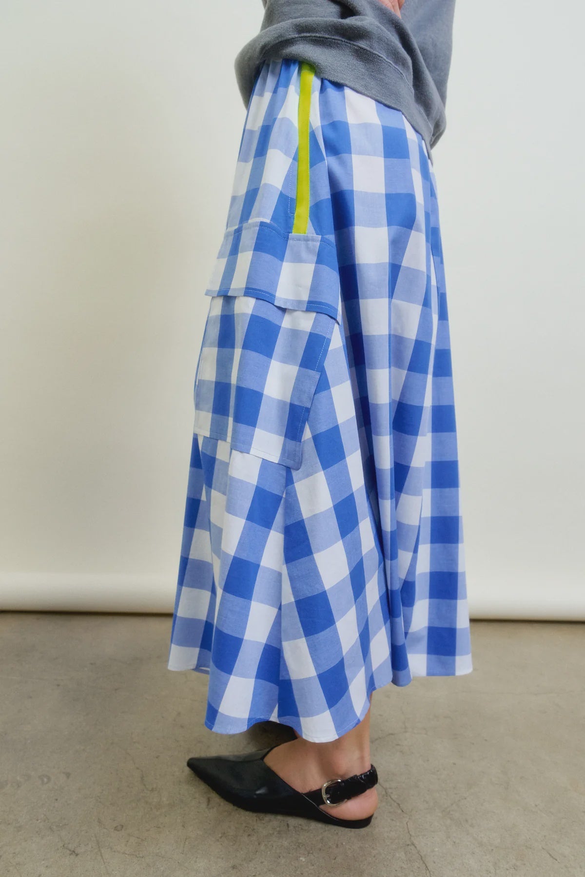 A person wears the Aquarius Cocktail Le Pencil Slim Skirt in blue and white Japanese cotton poplin with oversized cargo pockets and a yellow zipper, styled with black pointed-toe flats and a grey top, standing on a plain concrete floor.