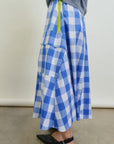 A person wears the Aquarius Cocktail Le Pencil Slim Skirt in blue and white Japanese cotton poplin with oversized cargo pockets and a yellow zipper, styled with black pointed-toe flats and a grey top, standing on a plain concrete floor.