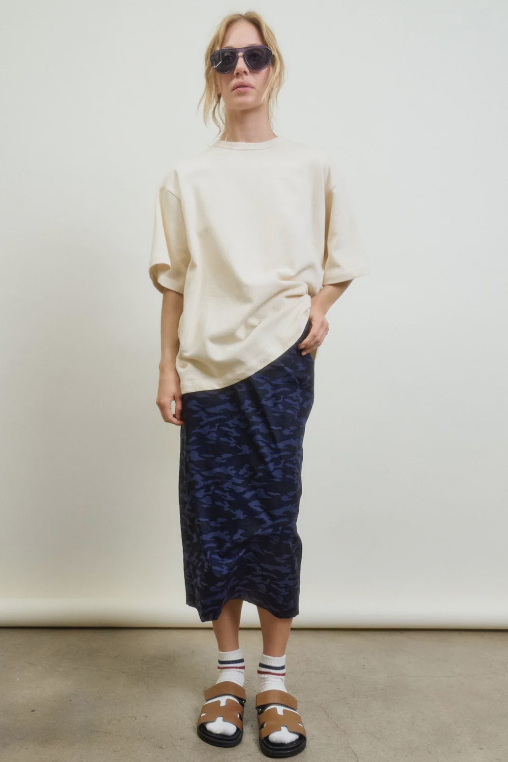 A woman in sunglasses, an oversized beige LA-made t-shirt, striped socks, brown sandals, and the Aquarius Cocktail CAMO LE Pencil Slim Skirt stands against a plain white background.