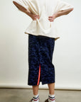 A person stands with hands on hips wearing the Aquarius Cocktail CAMO LE Pencil Slim Skirt in blue camo Japanese cotton, paired with a cream top, white socks, and black sandals. Photo taken from behind against a light background.