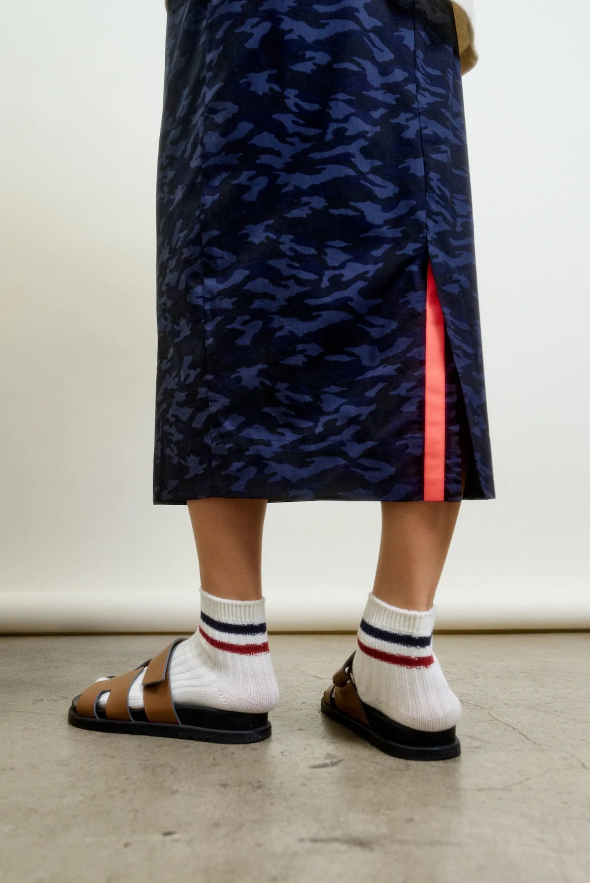 A person stands on concrete wearing the Aquarius Cocktail CAMO LE Pencil Slim Skirt in blue camo with a side slit and neon pink accent, paired with striped white socks and brown sandals.