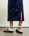A person stands on concrete wearing the Aquarius Cocktail CAMO LE Pencil Slim Skirt in blue camo with a side slit and neon pink accent, paired with striped white socks and brown sandals.