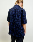 A person with light brown hair in a messy bun wears the Aquarius Cocktail CAMO LE Camp Shirt, featuring navy blue and black Japanese cotton, and stands facing away from the camera against a plain light background.