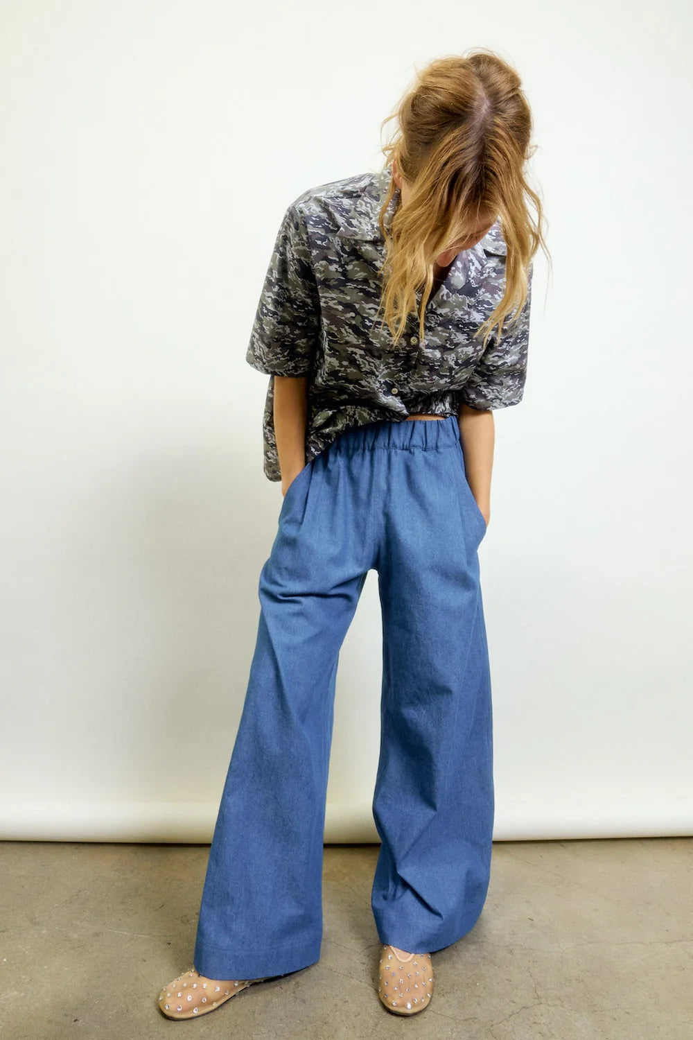 A person with long, light brown hair looks down, wearing a gray patterned shirt and Aquarius Cocktail’s Connor Wide Leg Pant in blue with an elastic waist, paired with beige shoes with white polka dots, standing against a plain light background.
