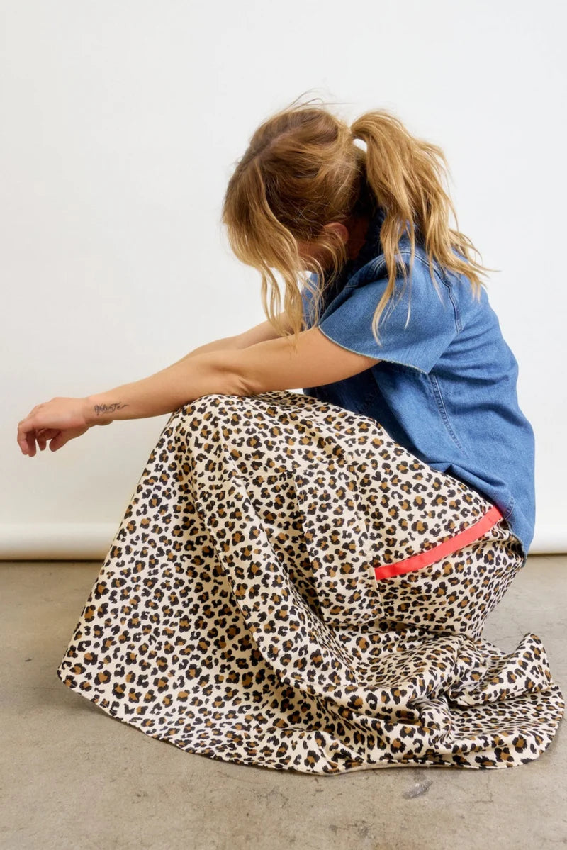 A person with light brown hair in a ponytail sits on the floor against a plain light background, wearing a blue short-sleeve shirt and the Aquarius Cocktail Le Cargo Skirt—a long leopard-print cargo skirt with oversized pockets and a pink stripe.
