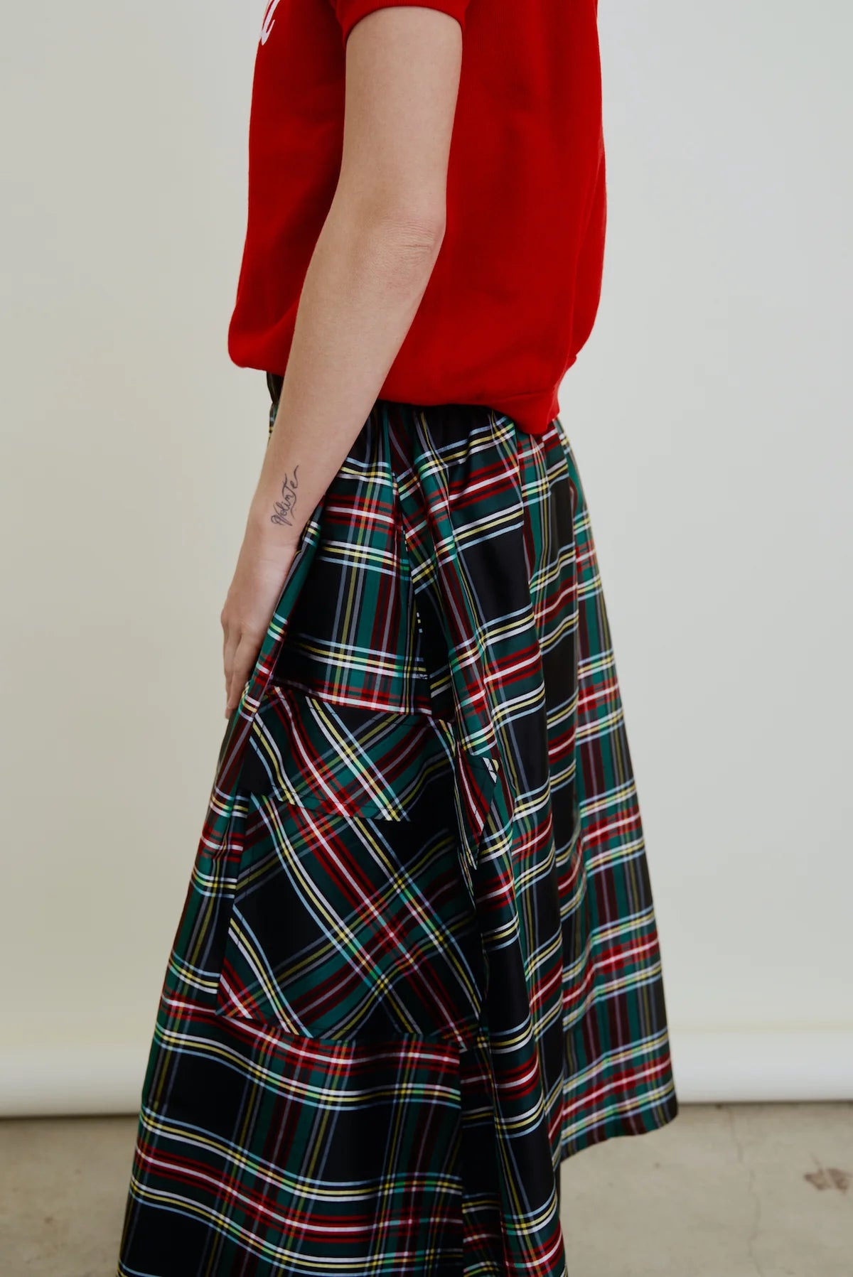 A person wears Aquarius Cocktail's Le Cargo Skirt—a long, black plaid piece with red, green, blue, and yellow stripes—paired with a red short-sleeve top. Their tattooed right arm rests at their side. Skirt is made in Los Angeles; background is light and plain.