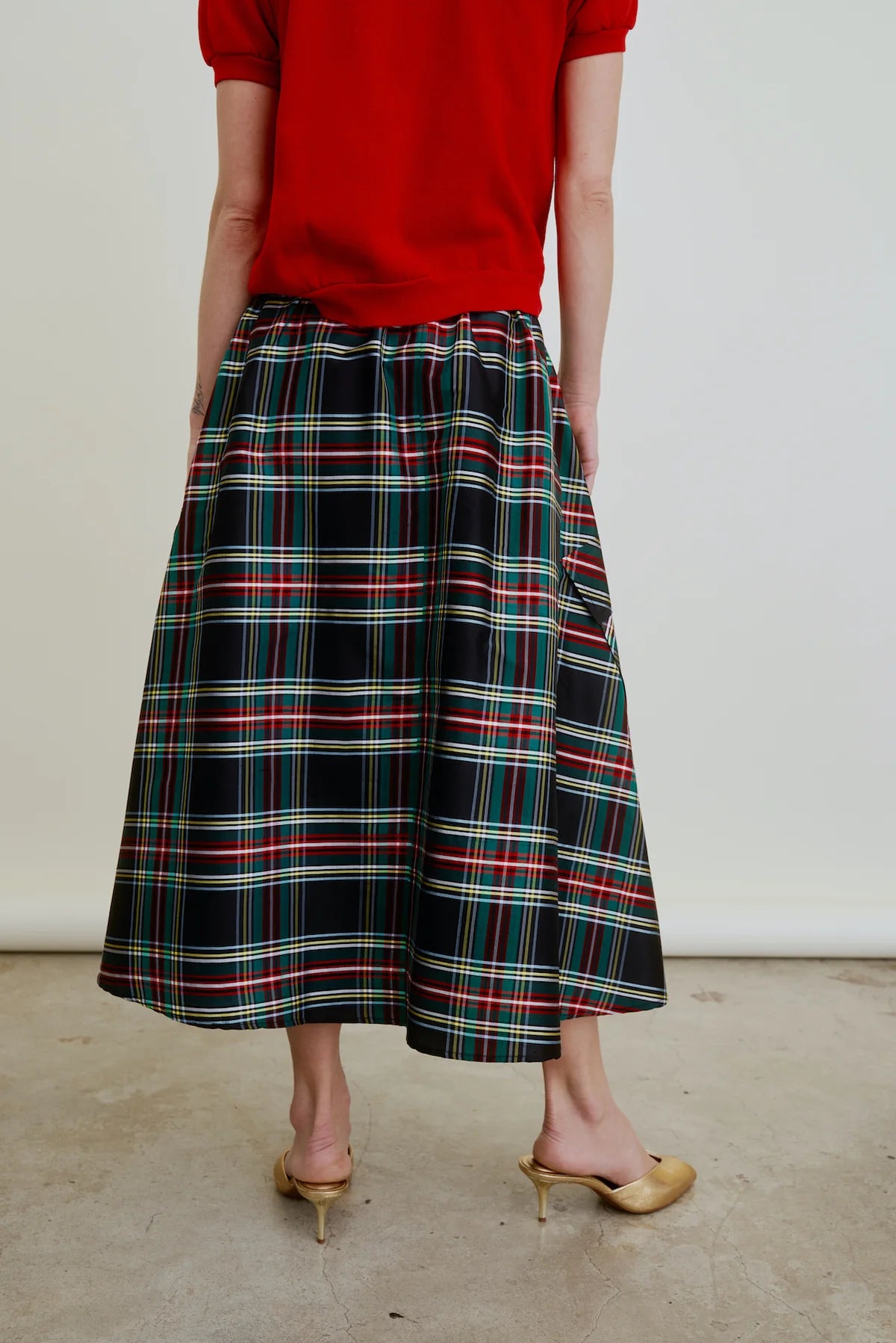 A person stands on a concrete floor wearing a red top, gold kitten heel shoes, and the Aquarius Cocktail Le Cargo Skirt—a black Japanese cotton poplin skirt with a colorful plaid pattern. The image is cropped from the shoulders down.
