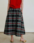 A person stands on a concrete floor wearing a red top, gold kitten heel shoes, and the Aquarius Cocktail Le Cargo Skirt—a black Japanese cotton poplin skirt with a colorful plaid pattern. The image is cropped from the shoulders down.