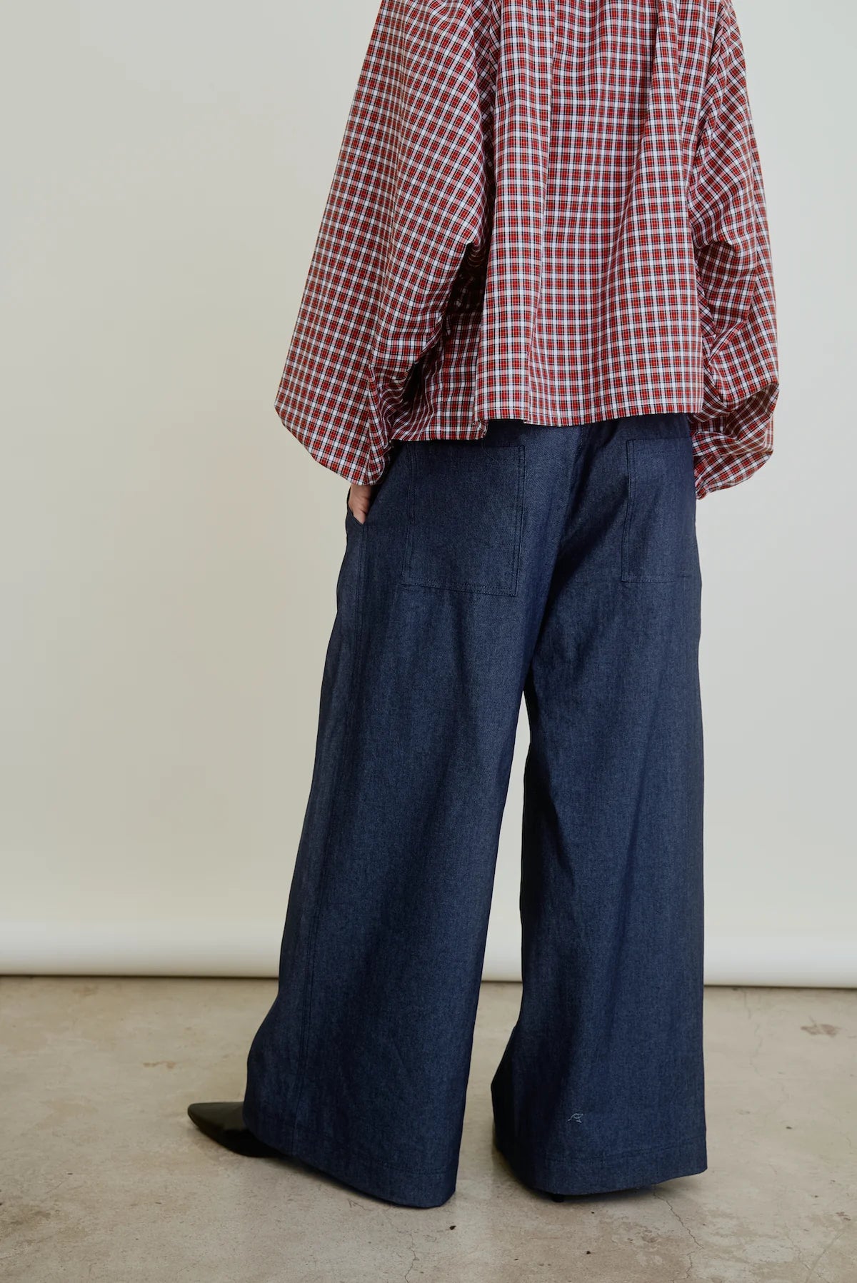 A person models the Connor Wide Leg Pant by Aquarius Cocktail indoors, wearing a red and white checkered long-sleeve shirt. Their hands are in their pockets, the background is plain, and the photo is cropped above the shoulders.